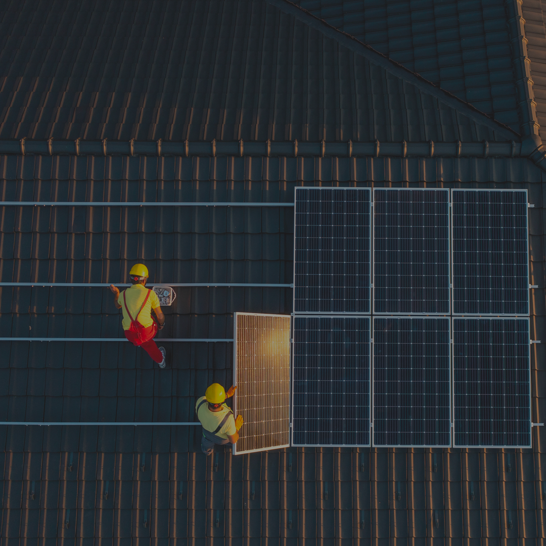 Operai ripresi dall'alto che montano pannelli fotovoltaici, sono vestiti con dei caschetti protettivi, maglietta e salopette.