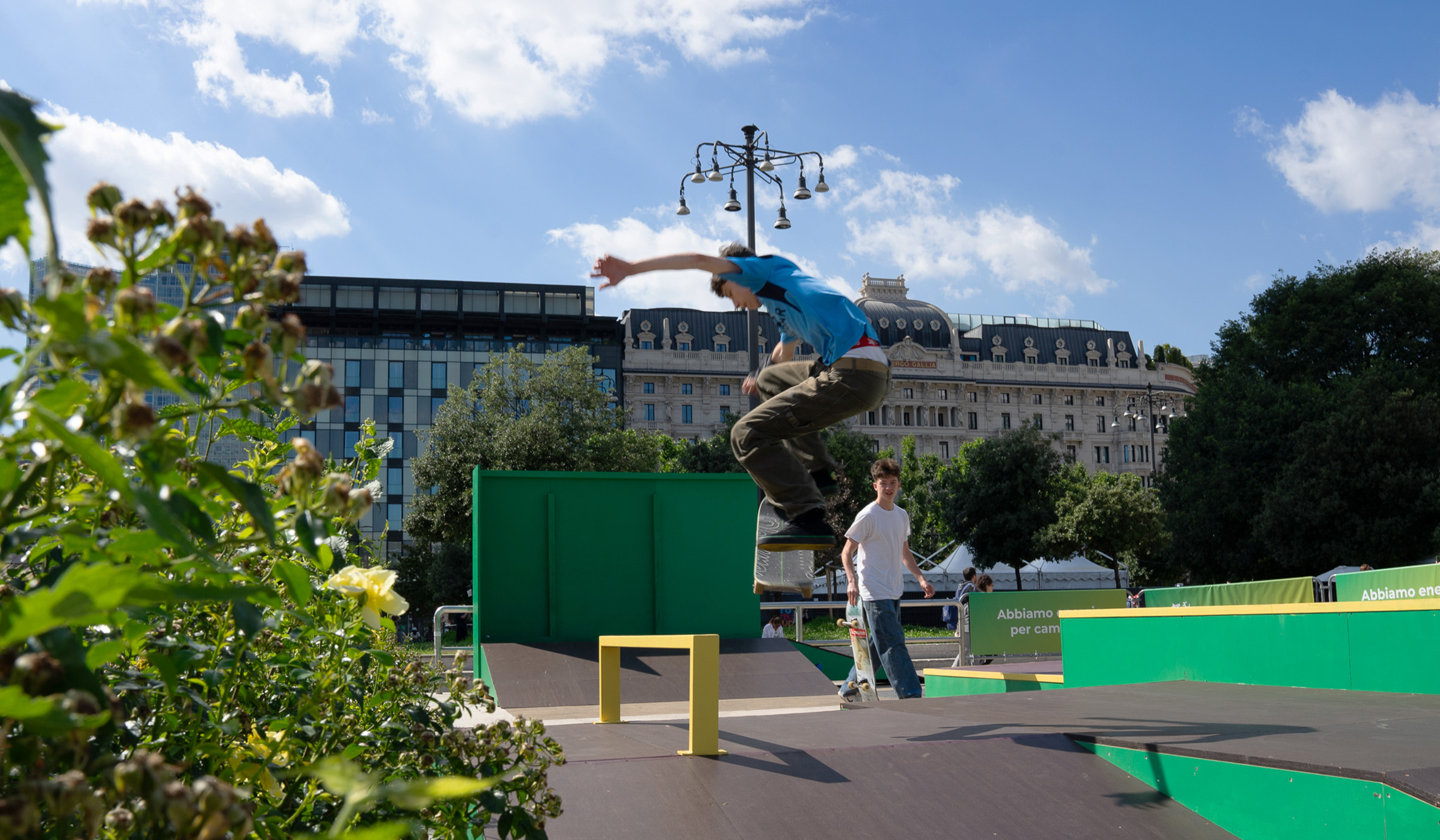 The Plenitude branded skater park with one boy on a skateboard and another looking on with skateboard in hand. In the background, Piazza Duca d'Aosta. 