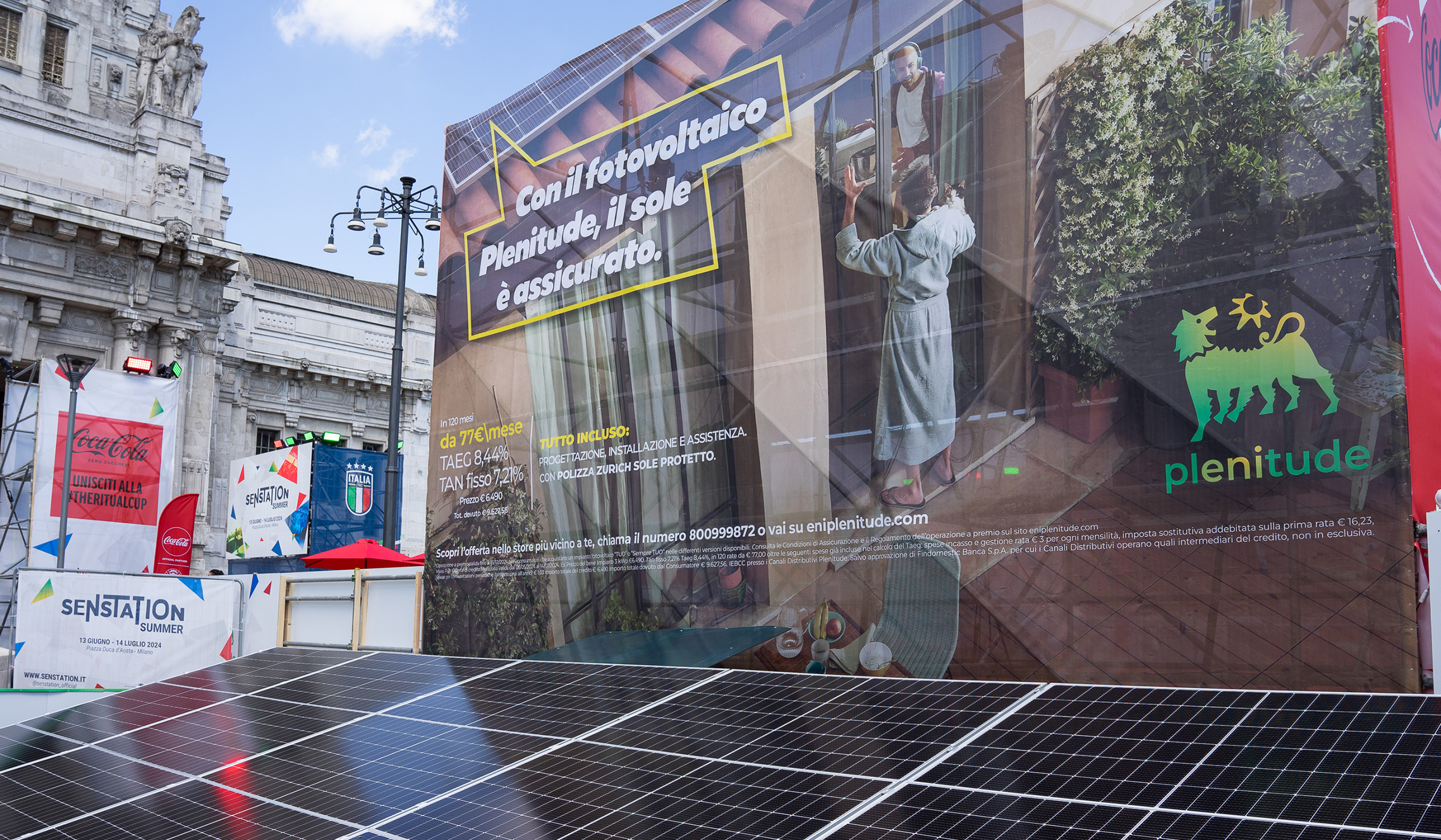 In the foreground are solar panels and a billboard with a picture of two boys, the first is inside the house and the second is outside the French window in a bathrobe, there is also the Plenitude logo and an inscription that says With Plenitude photovoltaics, the sun is assured. In the background, Milan's Central Station.  