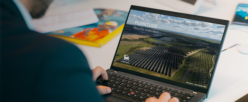 Un ordinateur portable ouvert posé sur une table, avec à l’écran l’inscription « Plenitude Results Overview », le logo d’Eni Plenitude et, en arrière-plan, un parc photovoltaïque.