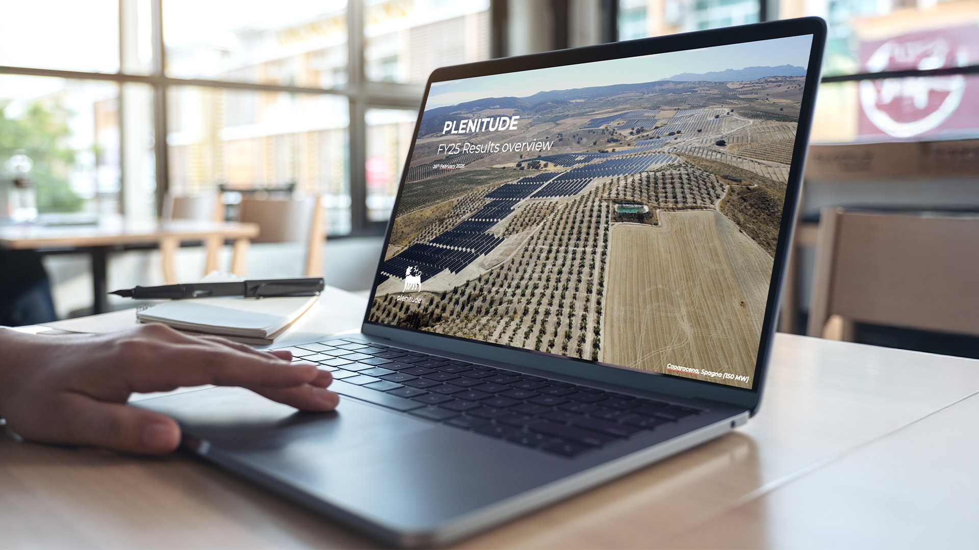 Um portátil aberto apoiado sobre uma mesa, no ecrã a inscrição “Plenitude Results Overview”, o logótipo da Eni Plenitude e, ao fundo, um parque fotovoltaico.